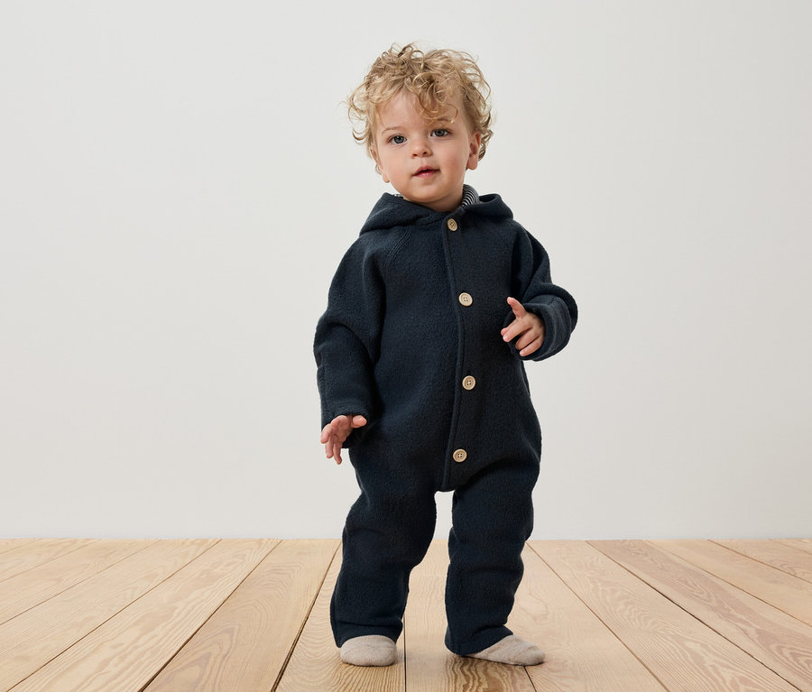 Un enfant aux cheveux bouclés se tient sur un plancher en bois, portant une combinaison bleu foncé à capuche et boutonnée.