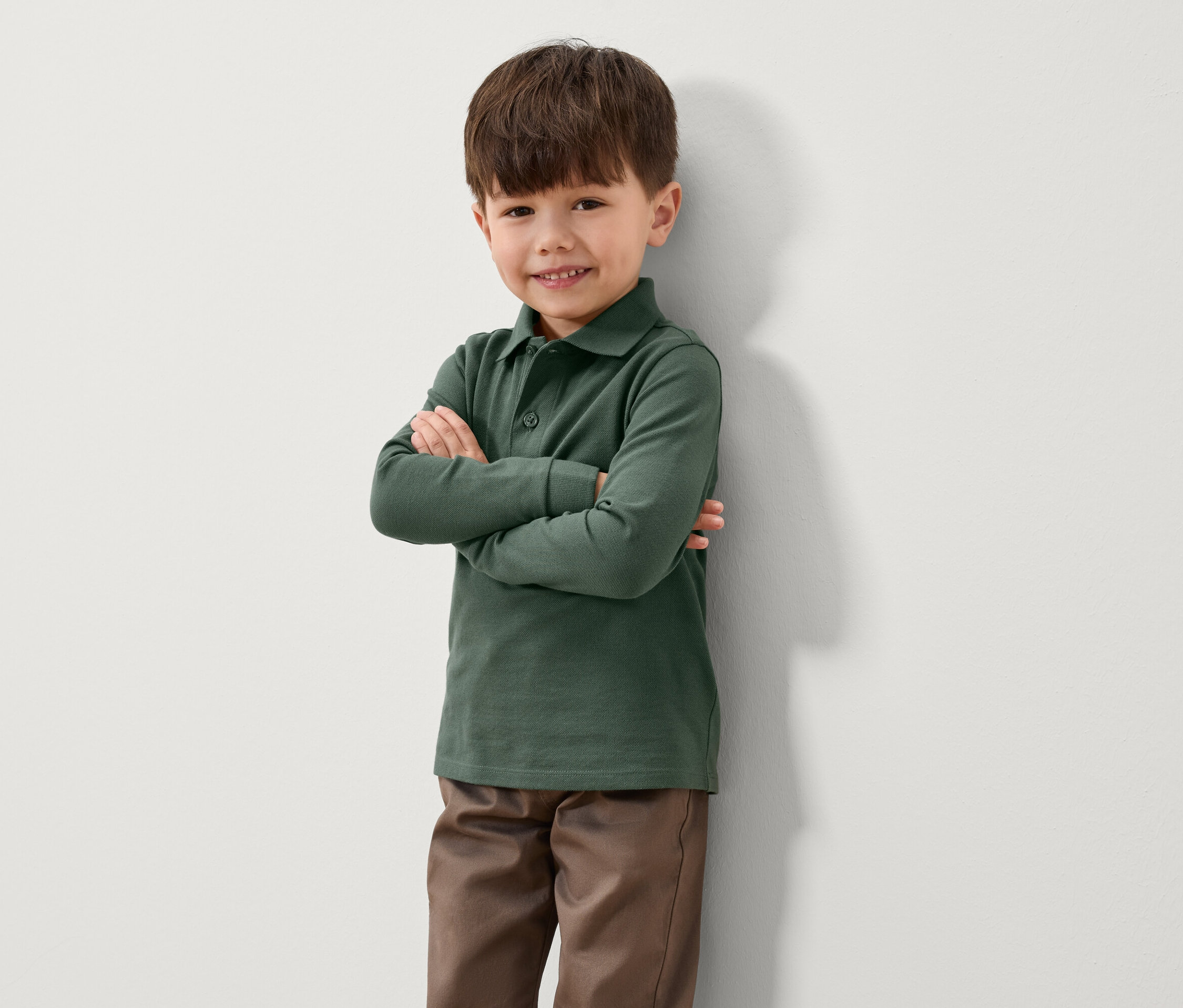 Un garçon se tient debout, les bras croisés, contre un mur. Il porte un polo vert à manches longues et un pantalon marron.
