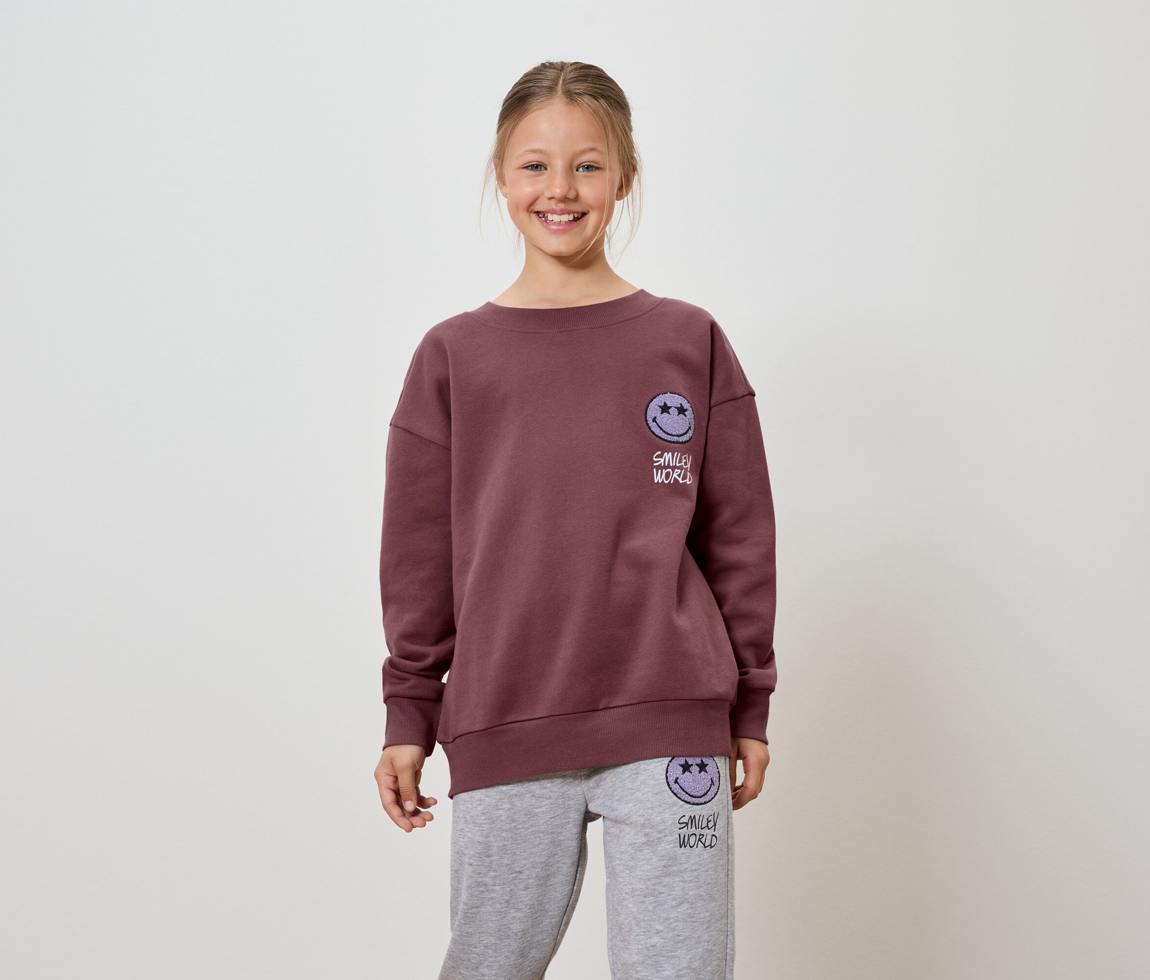 Une fillette souriante porte un sweat-shirt bordeaux et un pantalon de jogging gris avec un logo Smiley World.