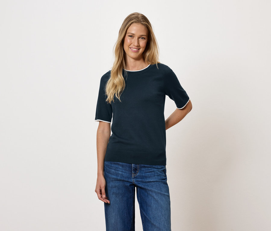 Une femme aux cheveux blonds porte un haut à manches courtes bleu foncé et un jean.