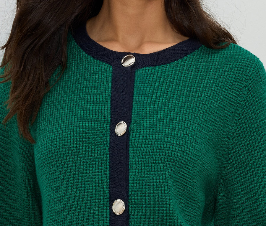 Gros plan d'une femme portant un cardigan en tricot vert avec des détails bleu marine et des boutons argentés.