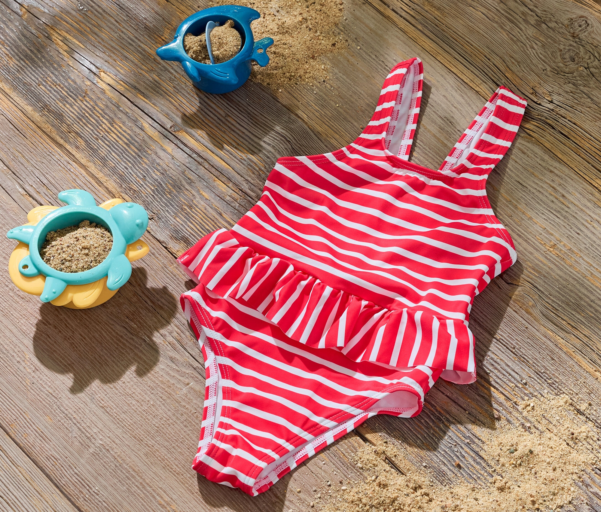 Un maillot de bain pour enfant rayé rouge et blanc est posé sur une surface en bois à côté de deux jouets de sable.