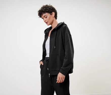 Une femme aux cheveux courts et bouclés porte un sweat à capuche noir, un pantalon de jogging noir et un débardeur blanc.