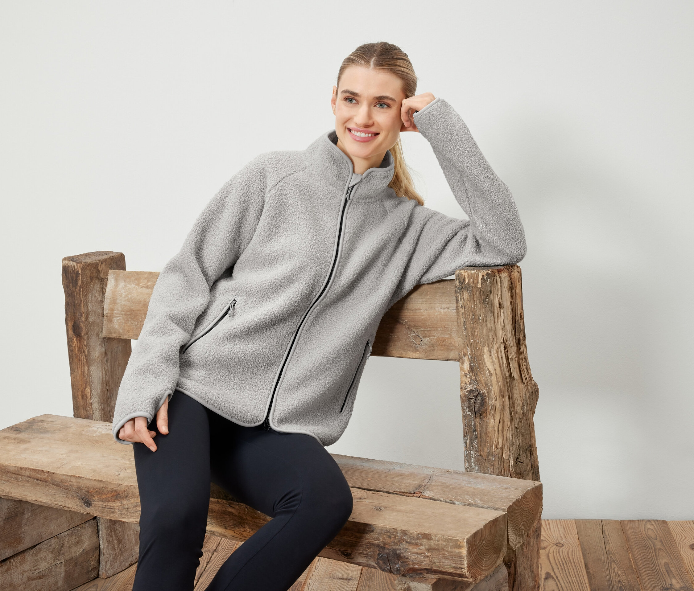 Femme en veste polaire grise posant sur un banc en bois.