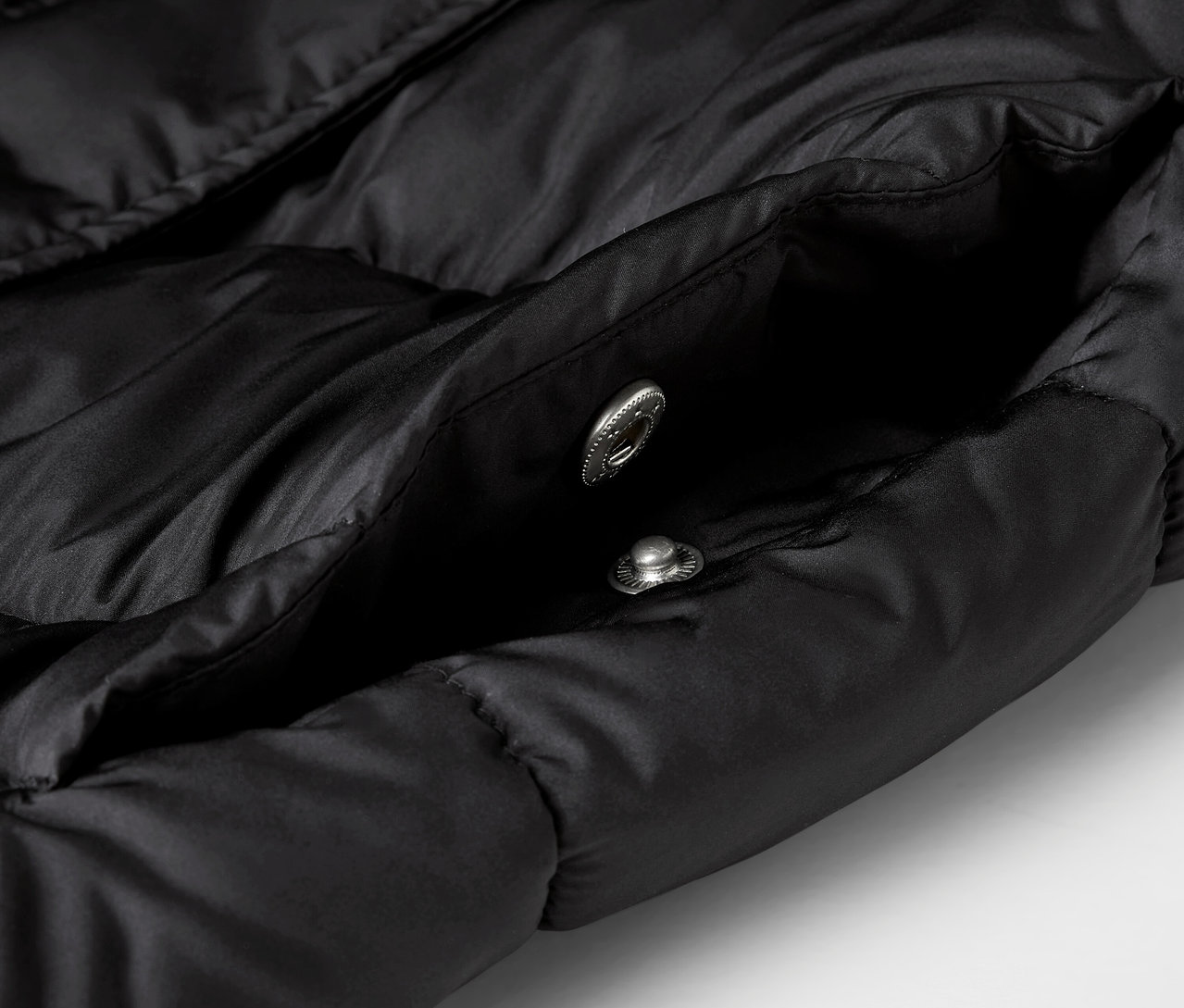 Gros plan sur une veste noire matelassée avec une poche ouverte et deux boutons-pression argentés.