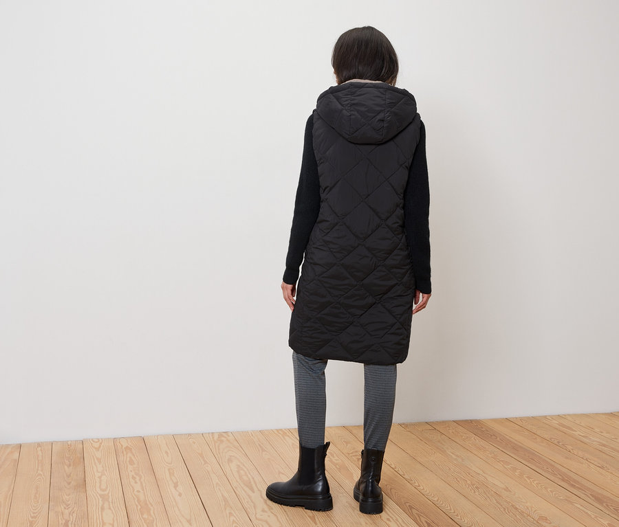 Une femme, de dos, porte une veste noire matelassée à capuche, des bottes noires et un legging gris.