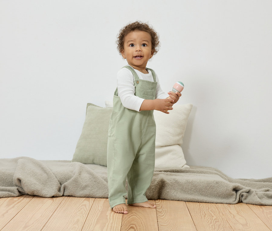 Un jeune enfant aux cheveux bouclés se tient sur un plancher en bois et tient un hochet. L'enfant porte une salopette verte sur un t-shirt blanc à manches longues.