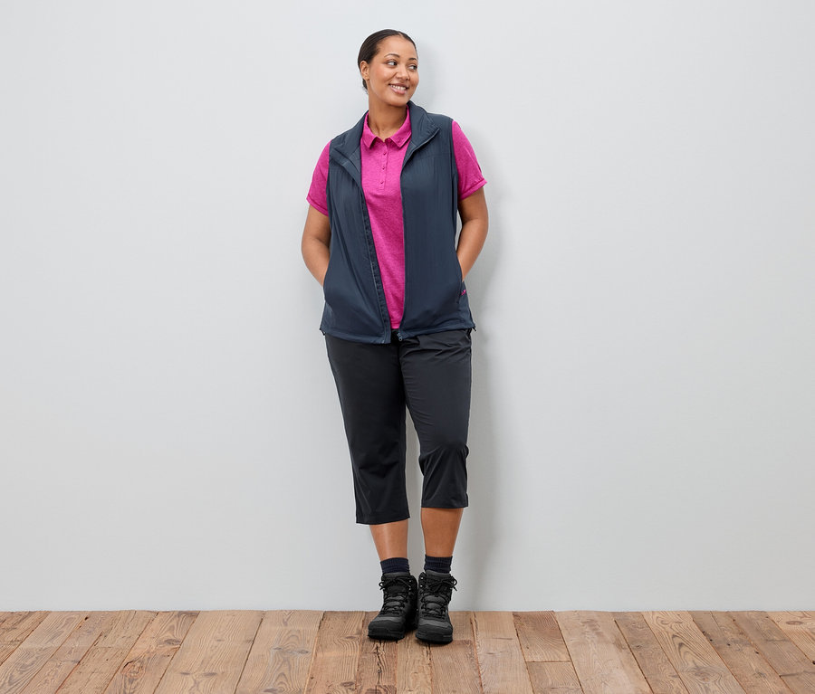 Une femme se tient devant un mur, les mains dans les poches. Elle porte une veste bleu marine sur un polo rose, avec un pantalon noir et des bottes.