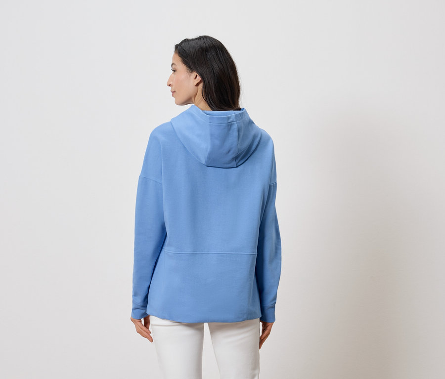 Femme vue de dos, portant un sweat à capuche bleu clair et un pantalon blanc sur un fond blanc.