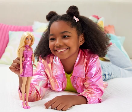 Une fille souriante, allongée sur le lit avec une veste rose, tient une poupée Barbie avec une robe rose.