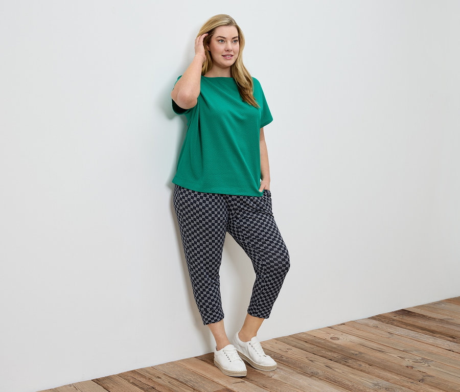Une femme en chemisier vert et pantalon à motifs est appuyée contre un mur blanc.