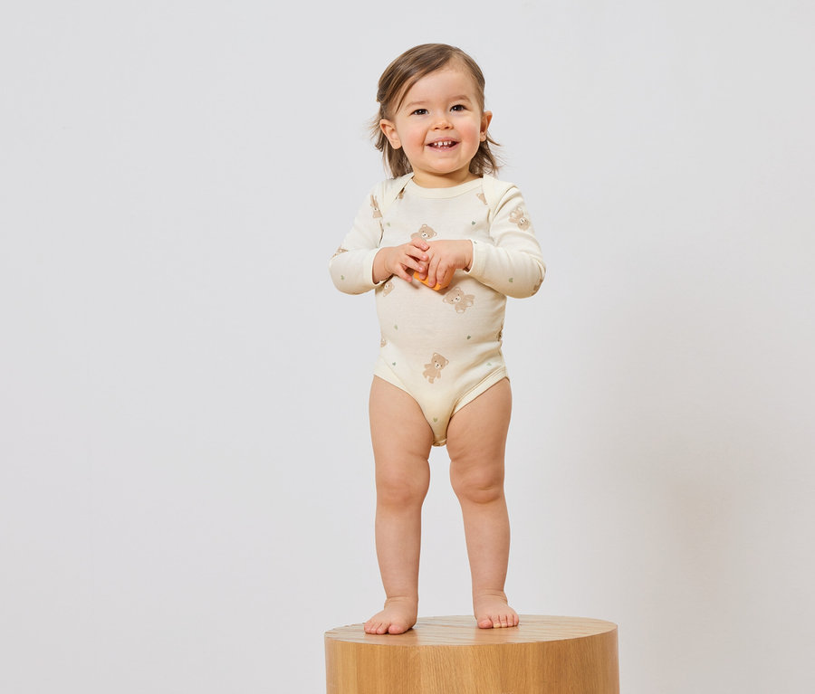 Un bébé souriant en body à motif d'ours en peluche se tient sur un tabouret devant un fond blanc.