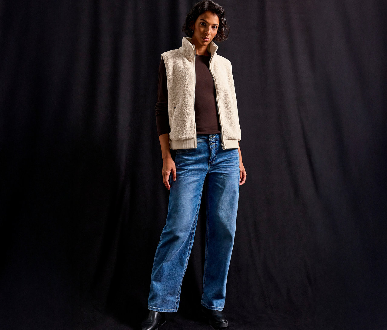 Une femme porte une veste en teddy Street One sur un t-shirt marron à manches longues et un jean sur un fond noir.