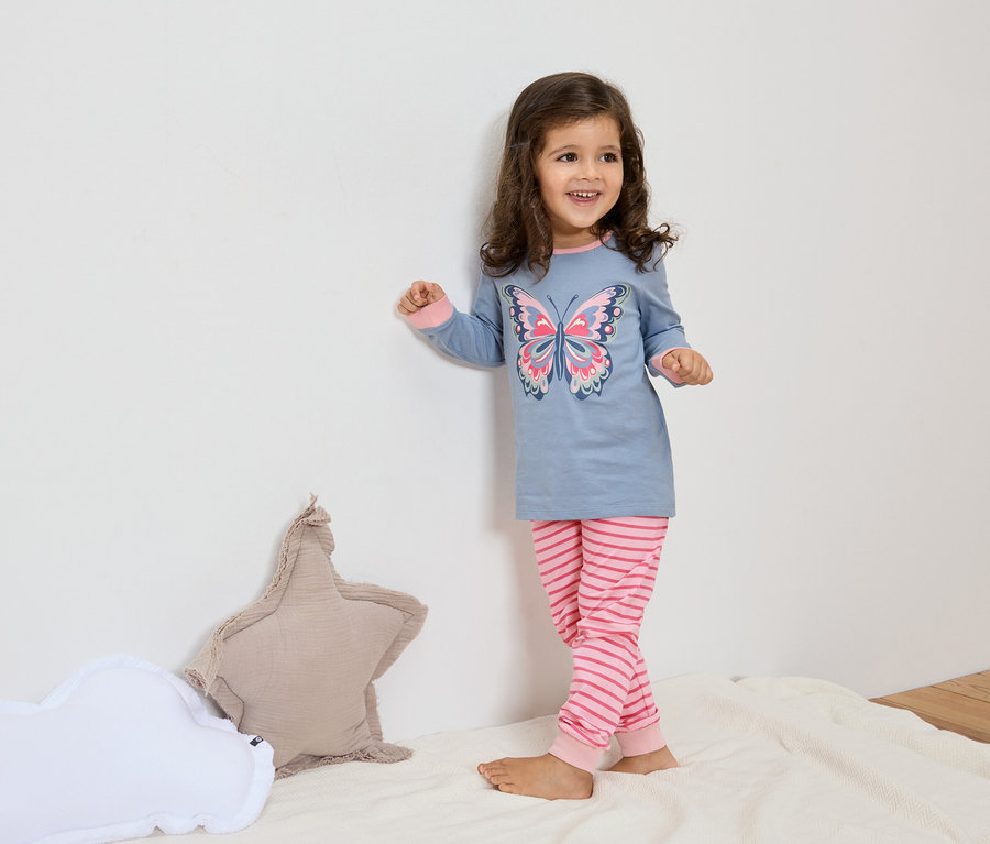 Une petite fille aux cheveux bruns porte un pyjama bleu avec un motif de papillon et un pantalon rayé rose. À côté d'elle se trouvent des coussins en forme d'étoile et de nuage sur une couverture blanche.
