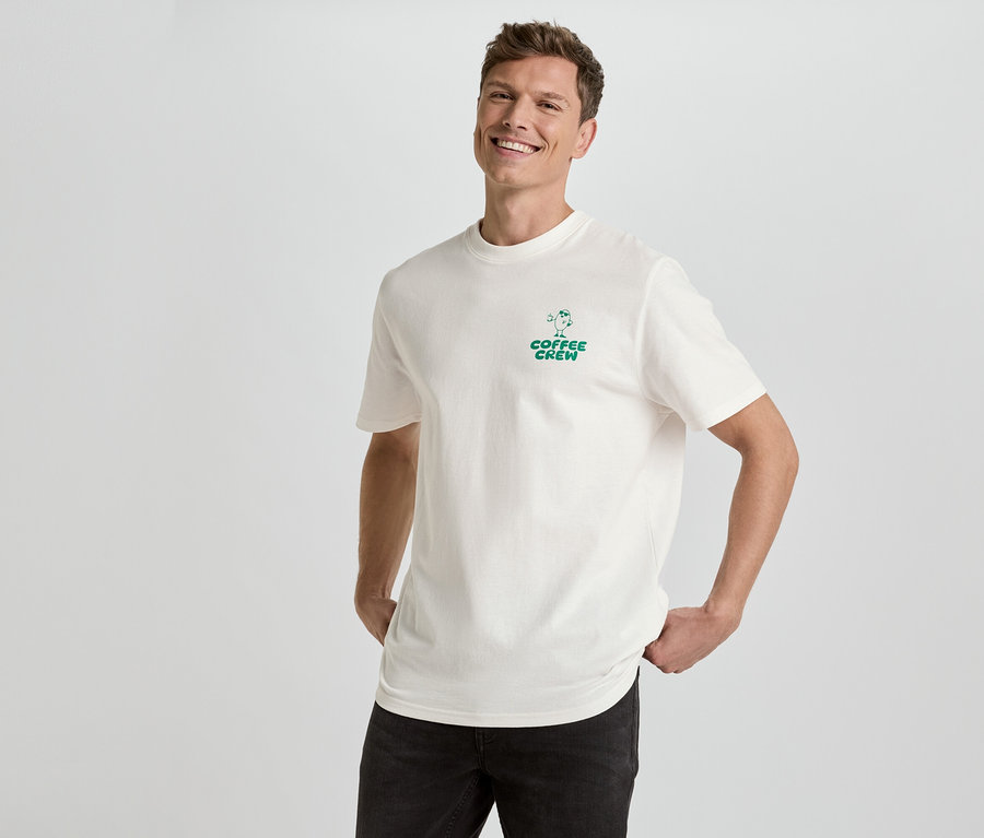 Un homme souriant pose en t-shirt crème avec un imprimé vert « Coffee Crew » et un jean noir sur fond blanc.