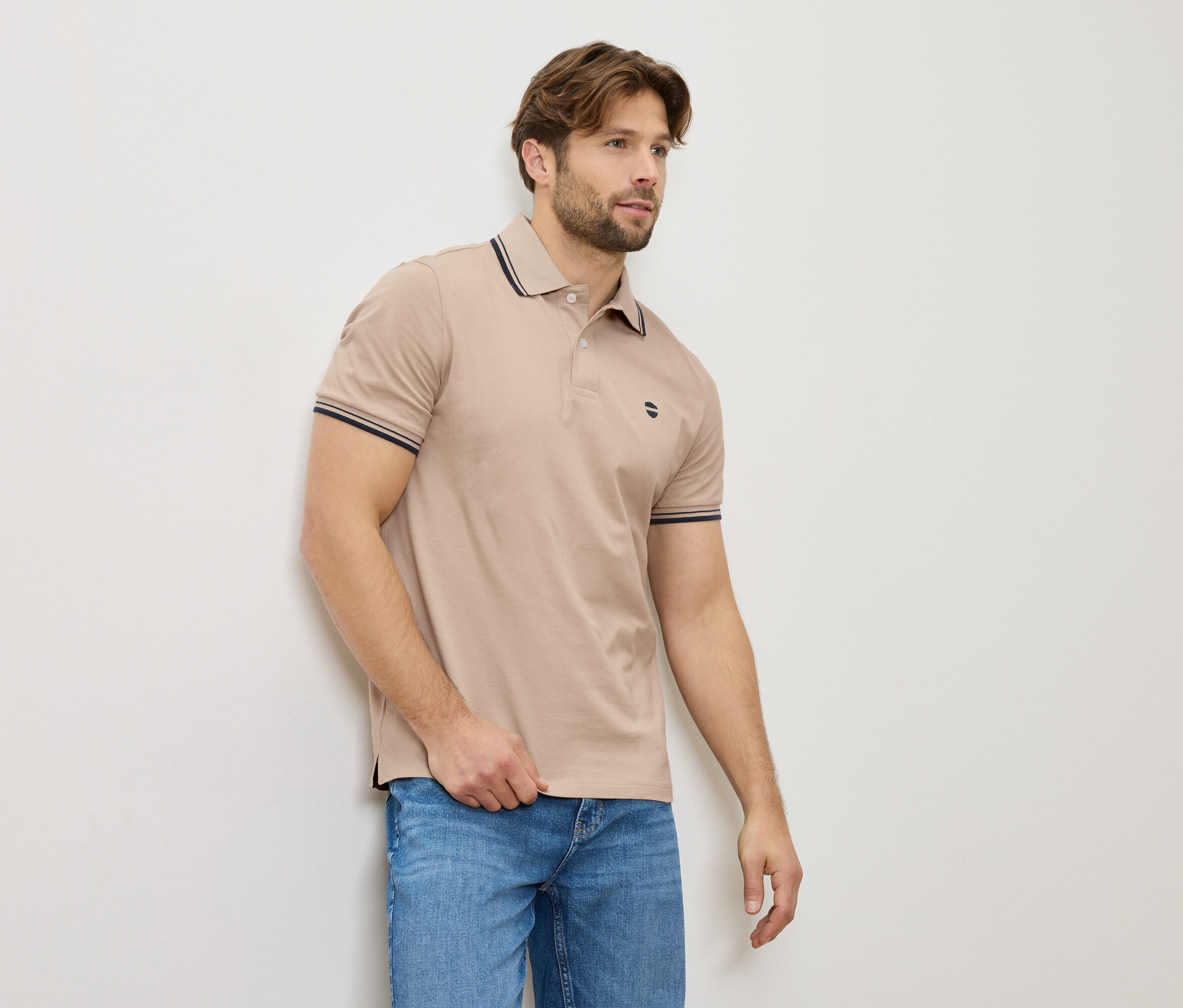Un homme en polo beige et jeans se tient contre un mur.