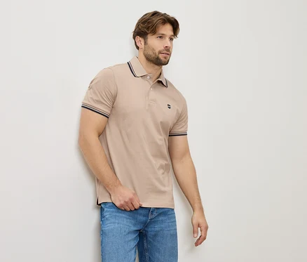 Un homme en polo beige et jeans se tient contre un mur.