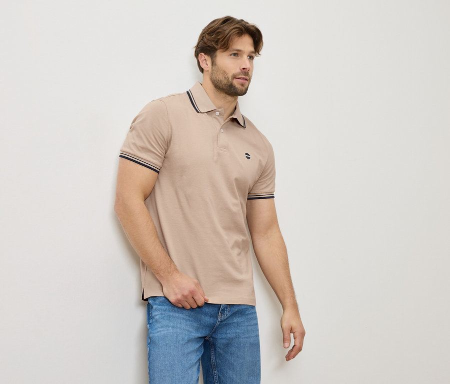 Un homme en polo beige et jeans se tient contre un mur.