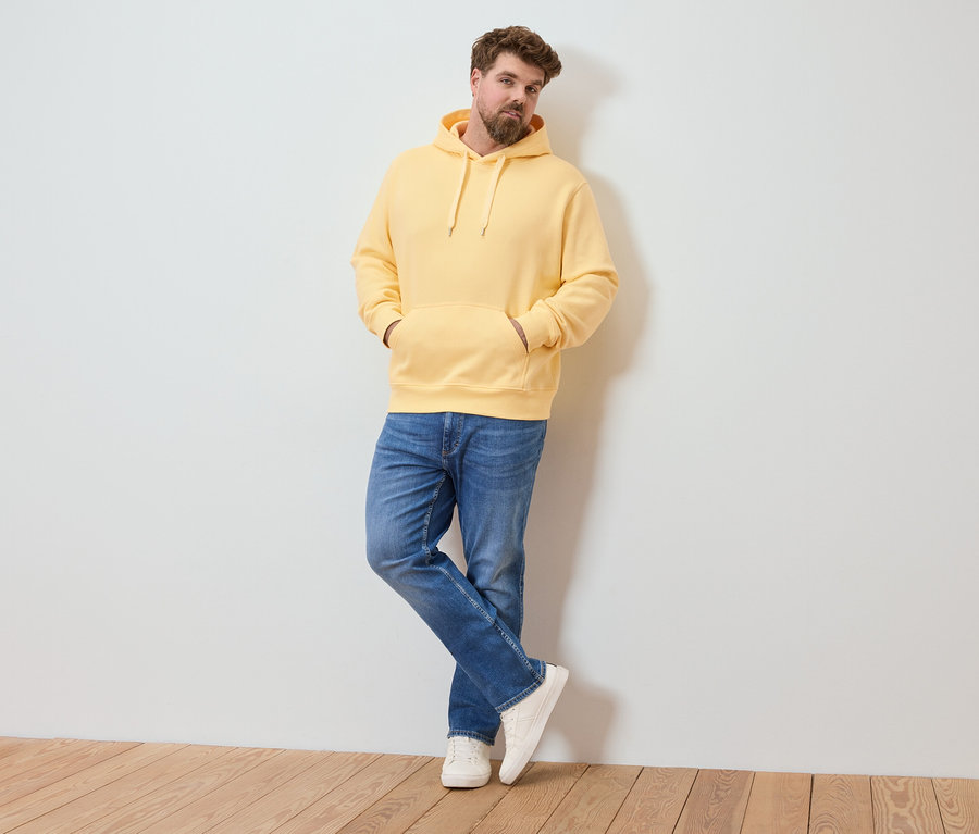 Un homme barbu est appuyé contre un mur, portant un sweat à capuche jaune, un jean et des baskets blanches.