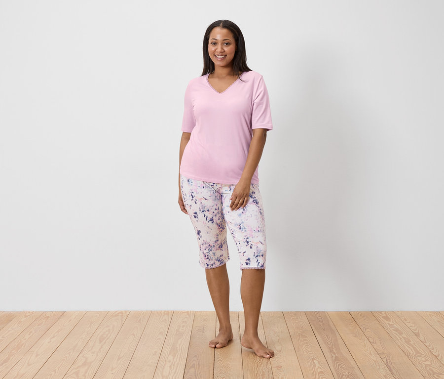 Femme en haut rose et pantalon fleuri debout pieds nus sur un plancher en bois devant un mur blanc.