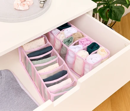 Une commode ouverte présente deux organiseurs en tissu rose à plusieurs compartiments remplis de vêtements roulés, et un t-shirt gris est partiellement visible.