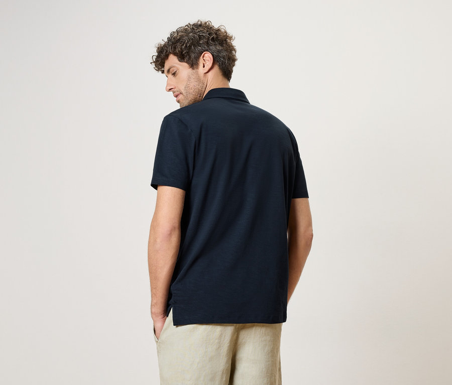 Un homme vu de dos, portant un polo bleu marine et un pantalon beige sur un fond neutre.