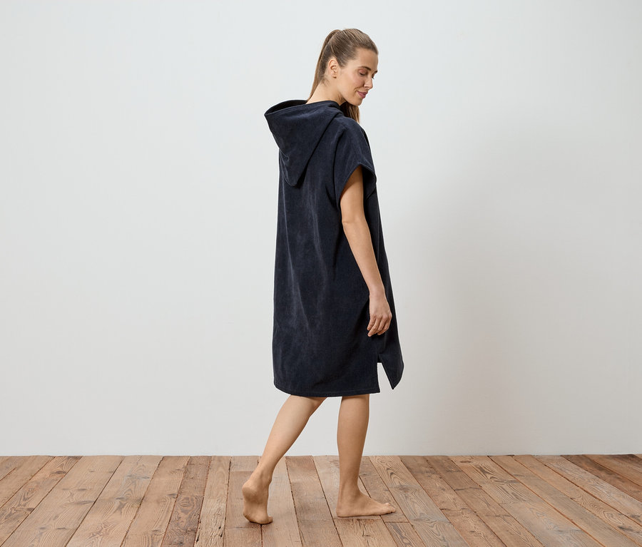Une femme pieds nus, portant un poncho à capuche bleu marine, marche sur un plancher en bois devant un mur blanc.
