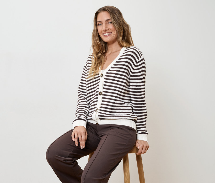 Une femme portant un pantalon marron et un cardigan rayé est assise sur un tabouret devant un fond blanc.