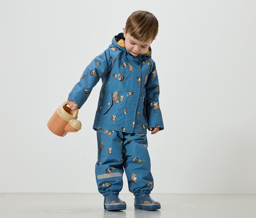 Un enfant arrose avec un arrosoir orange. Il porte un ensemble de pluie bleu à motifs d'animaux et des bottes en caoutchouc assorties.