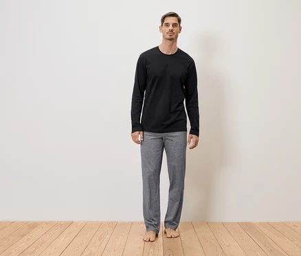 Un homme se tient pieds nus en pantalon gris et t-shirt noir à manches longues devant un mur sur un plancher en bois.
