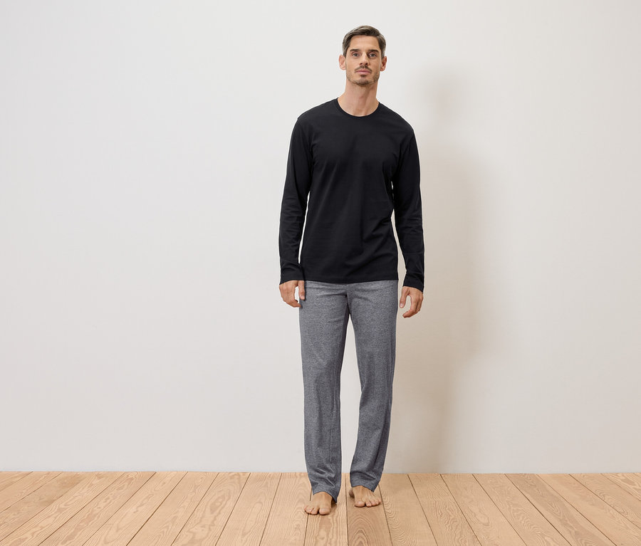 Un homme se tient pieds nus en pantalon gris et t-shirt noir à manches longues devant un mur sur un plancher en bois.