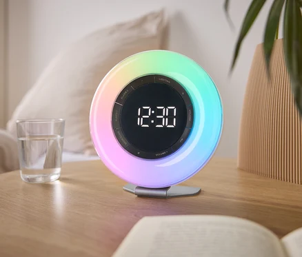 Sur une table de chevet se trouvent un radio-réveil lumineux coloré affichant 12h30, un verre d'eau et un livre.