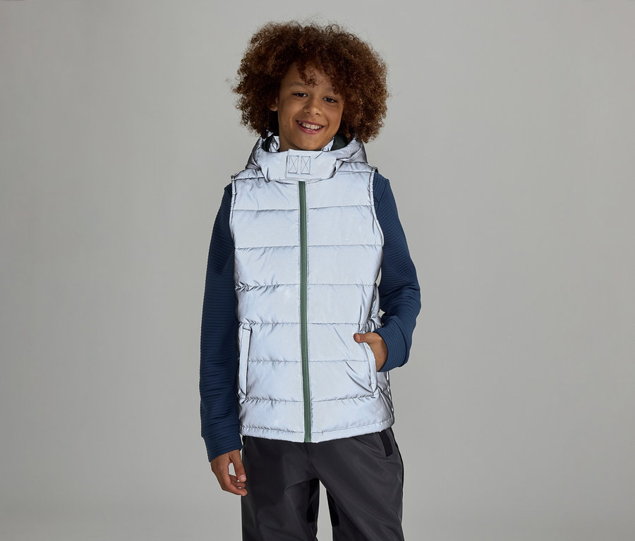 Un garçon avec un afro se tient sur un fond gris dans un gilet réfléchissant à capuche sur un pull bleu et un pantalon gris.