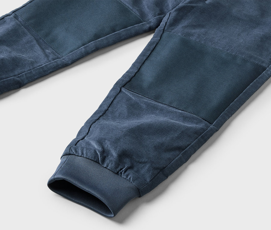 Gros plan d'un pantalon en velours côtelé bleu marine avec des poignets élastiques et des genoux renforcés sur un fond gris clair.