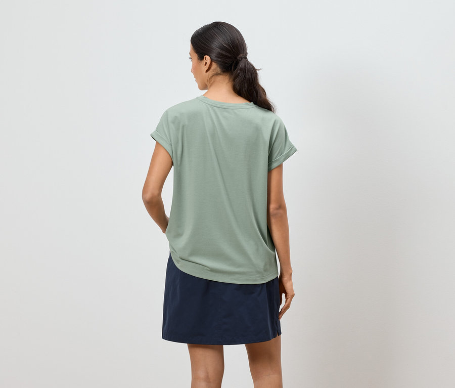 Femme de dos portant un t-shirt vert olive et une jupe bleue sur fond blanc.