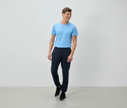 Un homme souriant se tient devant un mur blanc, vêtu d'un t-shirt bleu clair, d'un pantalon de jogging bleu marine et de chaussures de sport noires.