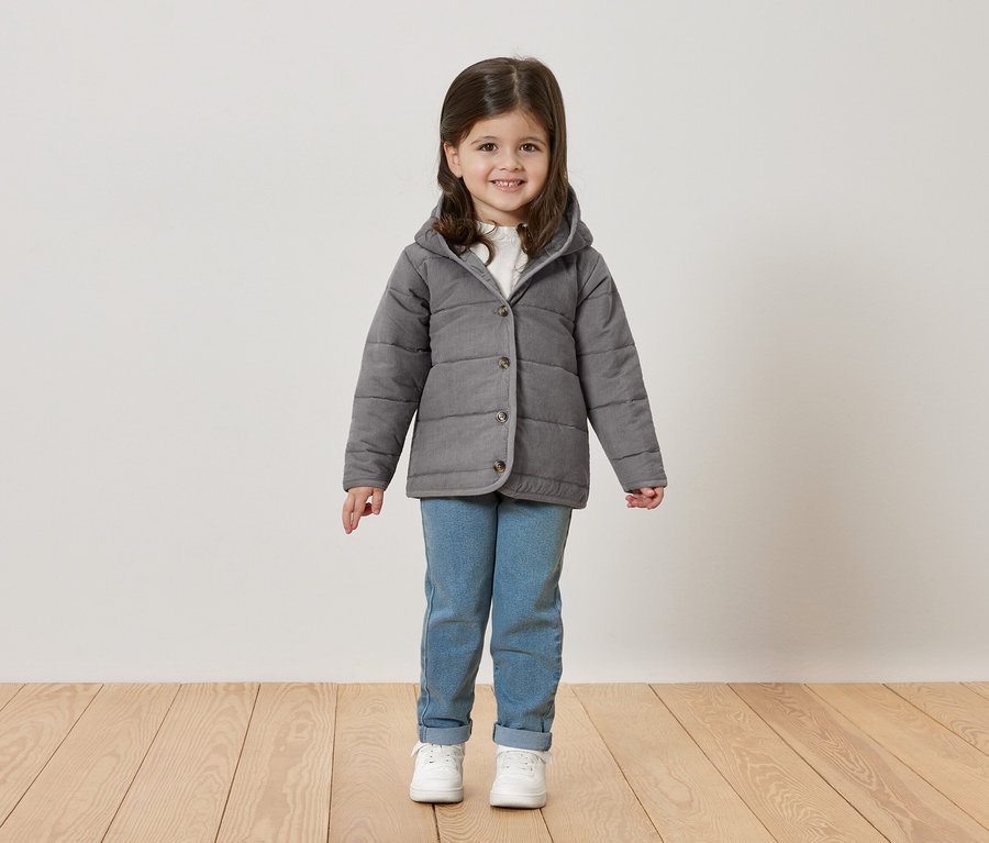 Une petite fille aux cheveux bruns sourit à la caméra, portant une veste grise, un jean et des baskets blanches.