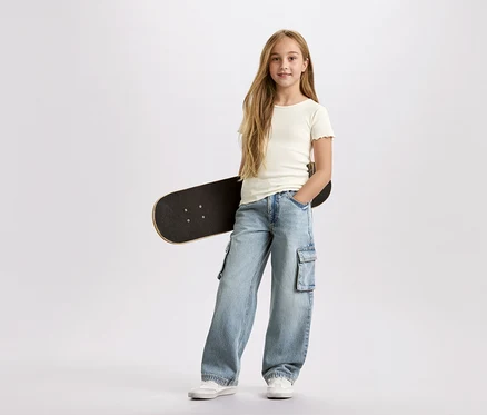 Une fille aux longs cheveux blonds, avec un skateboard noir sous le bras, porte un pantalon cargo bleu clair et un t-shirt blanc sur un fond blanc.