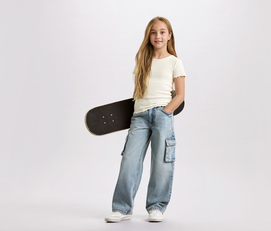 Une fille aux longs cheveux blonds, avec un skateboard noir sous le bras, porte un pantalon cargo bleu clair et un t-shirt blanc sur un fond blanc.