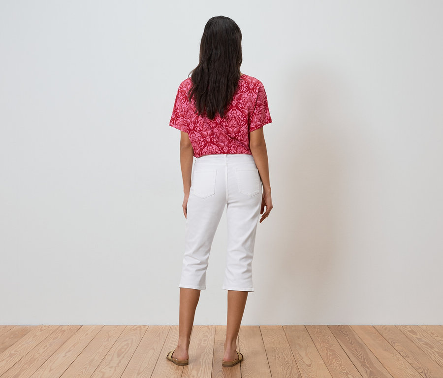 Une femme aux cheveux foncés se tient dos à la caméra, portant un haut à motifs rouges, un pantacourt blanc et des sandales dorées.