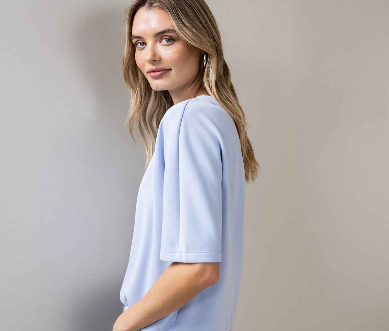 Une femme regarde par-dessus son épaule et porte une chemise Street One bleu clair à manches courtes et des créoles argentées.
