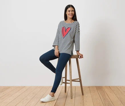 Une femme est assise sur un tabouret, portant un t-shirt rayé à manches longues avec un cœur, un legging et des baskets blanches.