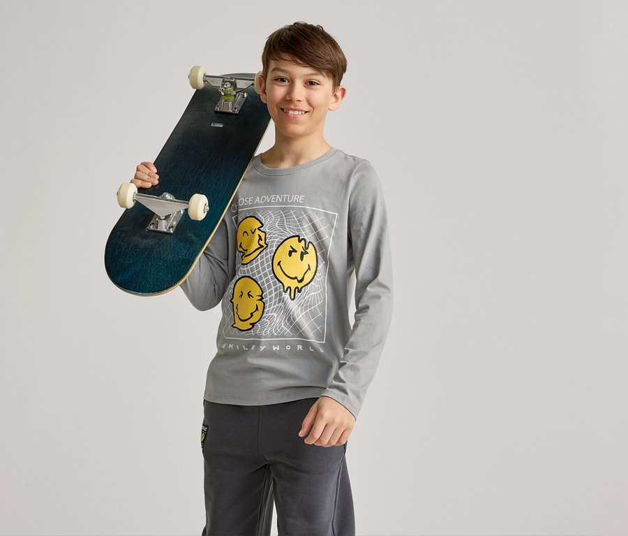 Un garçon souriant tient un skateboard sur son épaule, porte un t-shirt gris à manches longues avec des motifs de smileys jaunes et un pantalon gris.