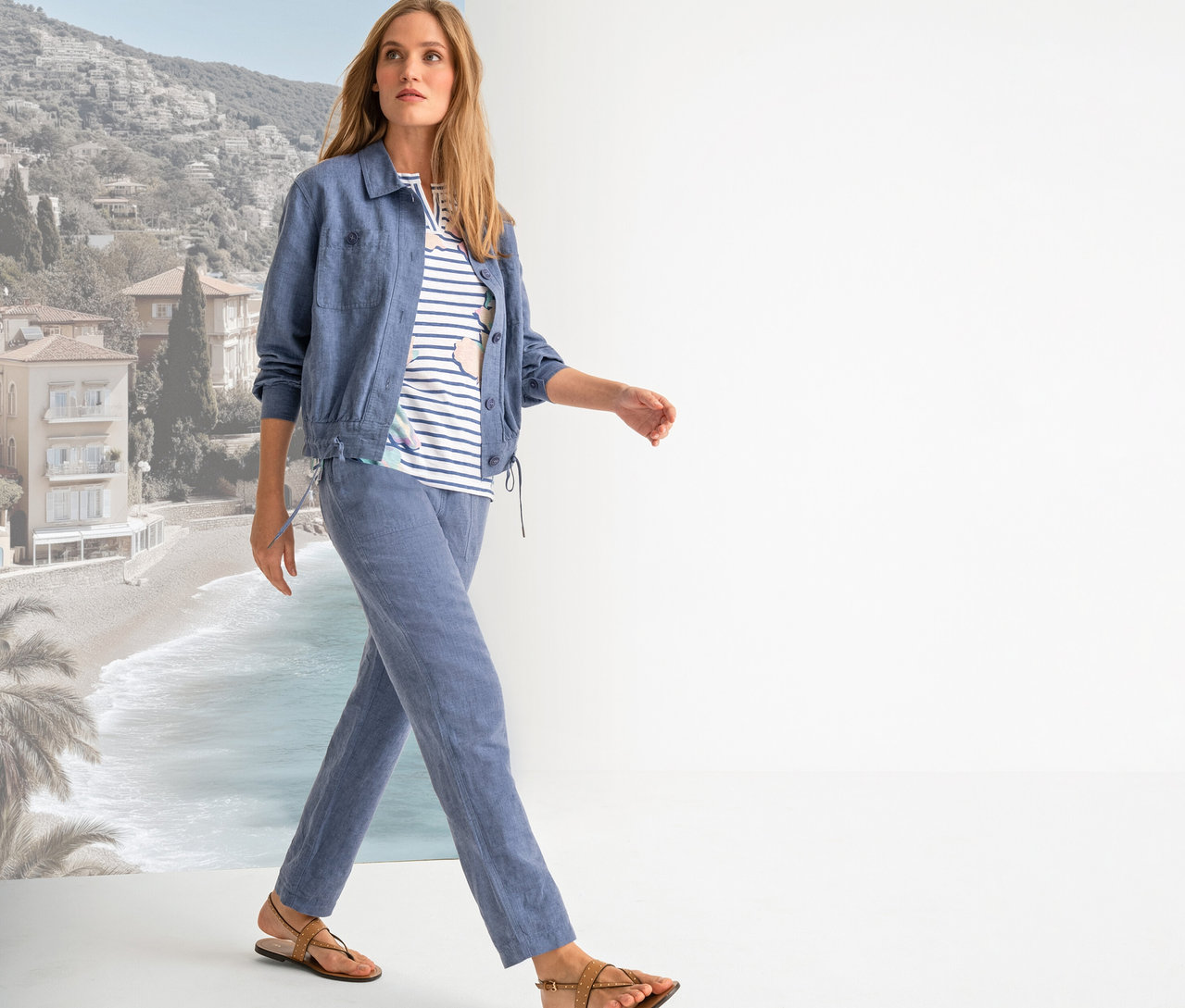 Une femme marche avec une veste bleue, un t-shirt rayé, un pantalon bleu et des sandales marron sur fond de plage.