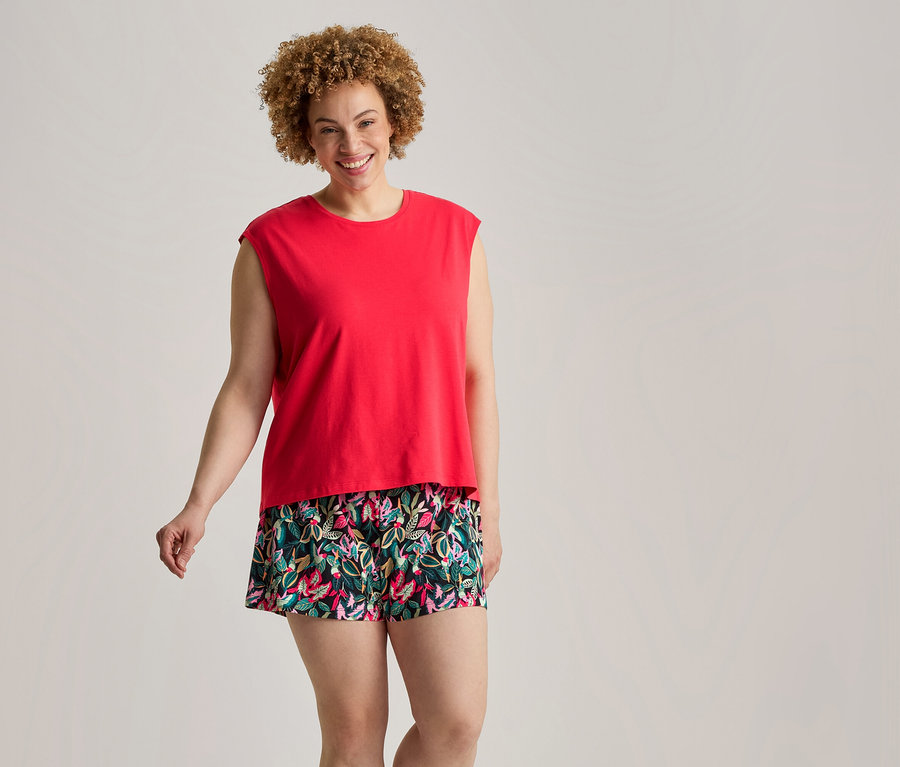 Femme souriante aux cheveux bouclés portant un haut rouge sans manches et un short court à motifs floraux verts, roses et bleus.