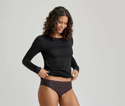 Une femme souriante aux cheveux bouclés, vêtue d'un haut noir à manches longues et d'un slip à motifs, ajustant son vêtement.