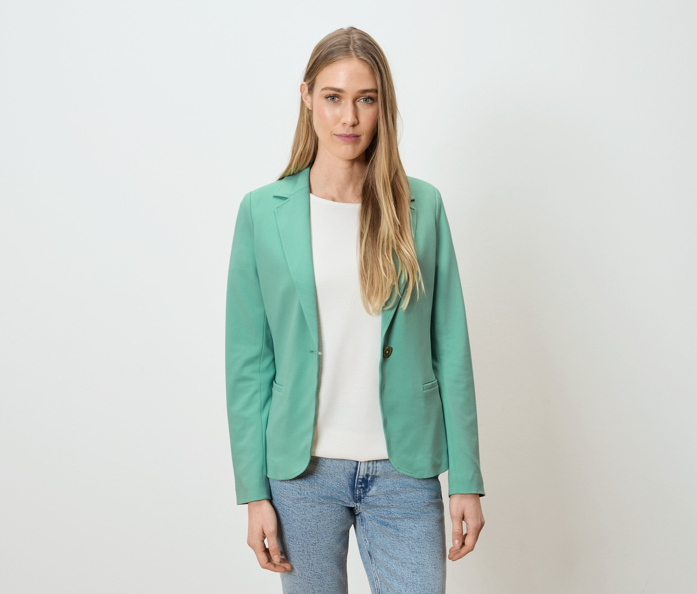 Une femme porte un blazer vert, un t-shirt blanc et un jean bleu sur un fond blanc.