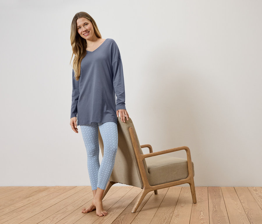 Une femme portant un long haut bleu et des leggings bleu clair à motifs, appuyée contre une chaise avec une assise beige.