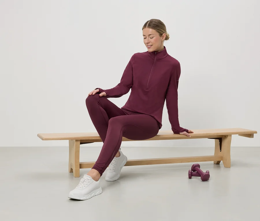 Une femme assise sur un banc porte une tenue de sport bordeaux avec un haut zippé, des leggings et des baskets blanches. Des haltères sont au sol.