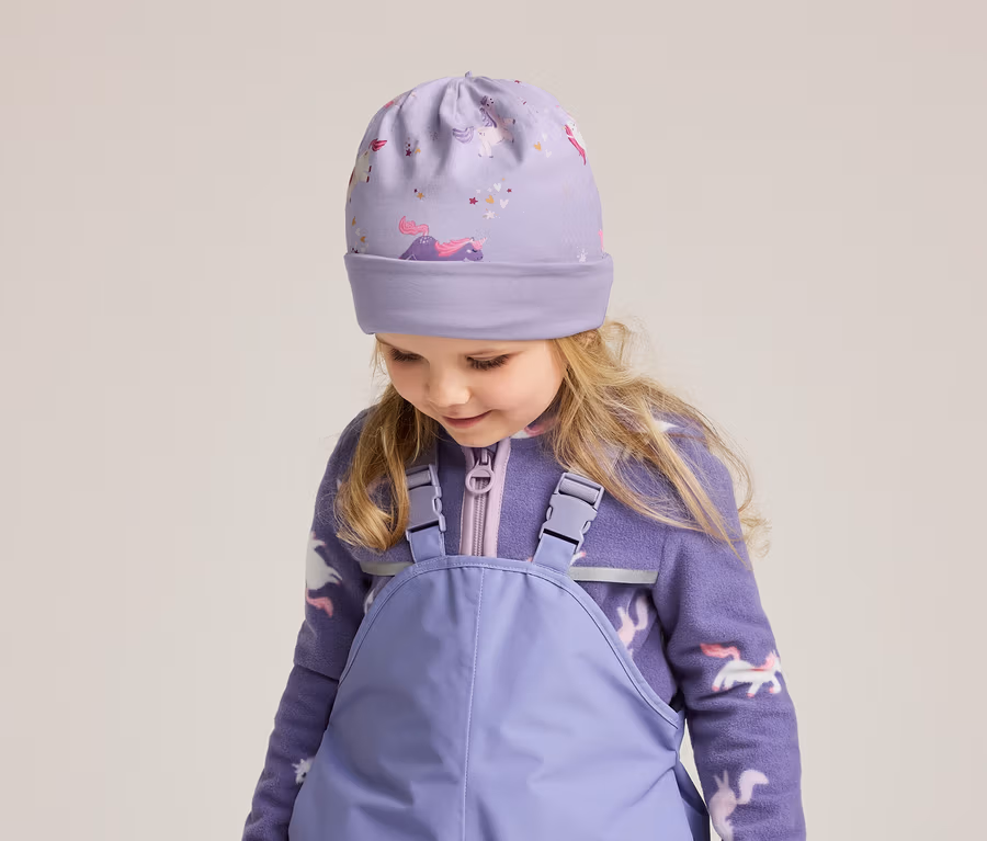 Une fillette blonde porte un bonnet violet à motifs de licornes, une veste polaire zippée et une salopette violette.